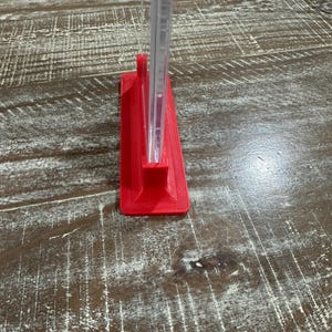 May include: A red, 3D-printed stand holding a clear, rectangular object. The stand is a solid, vibrant red color and has a rectangular base. The clear object appears to be a thin, transparent sheet or panel.