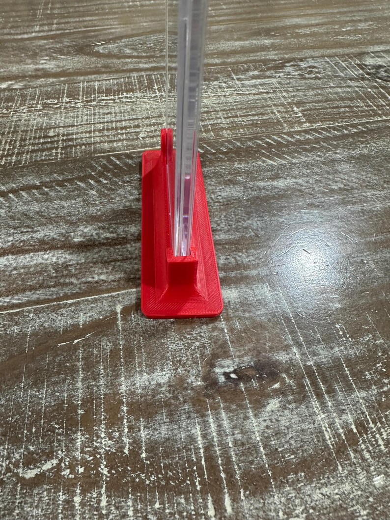 May include: A red, 3D-printed stand holding a clear, rectangular object. The stand is a solid, vibrant red color and has a rectangular base. The clear object appears to be a thin, transparent sheet or panel.