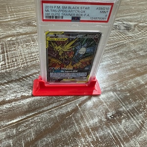 May include: A graded Pok&eacute;mon card in a clear plastic case, displayed on a red stand. The card features a colorful illustration of Moltres & Zapdos & Articuno GX. Text on the case indicates it's a 2019 P.M. SM Black Star card.