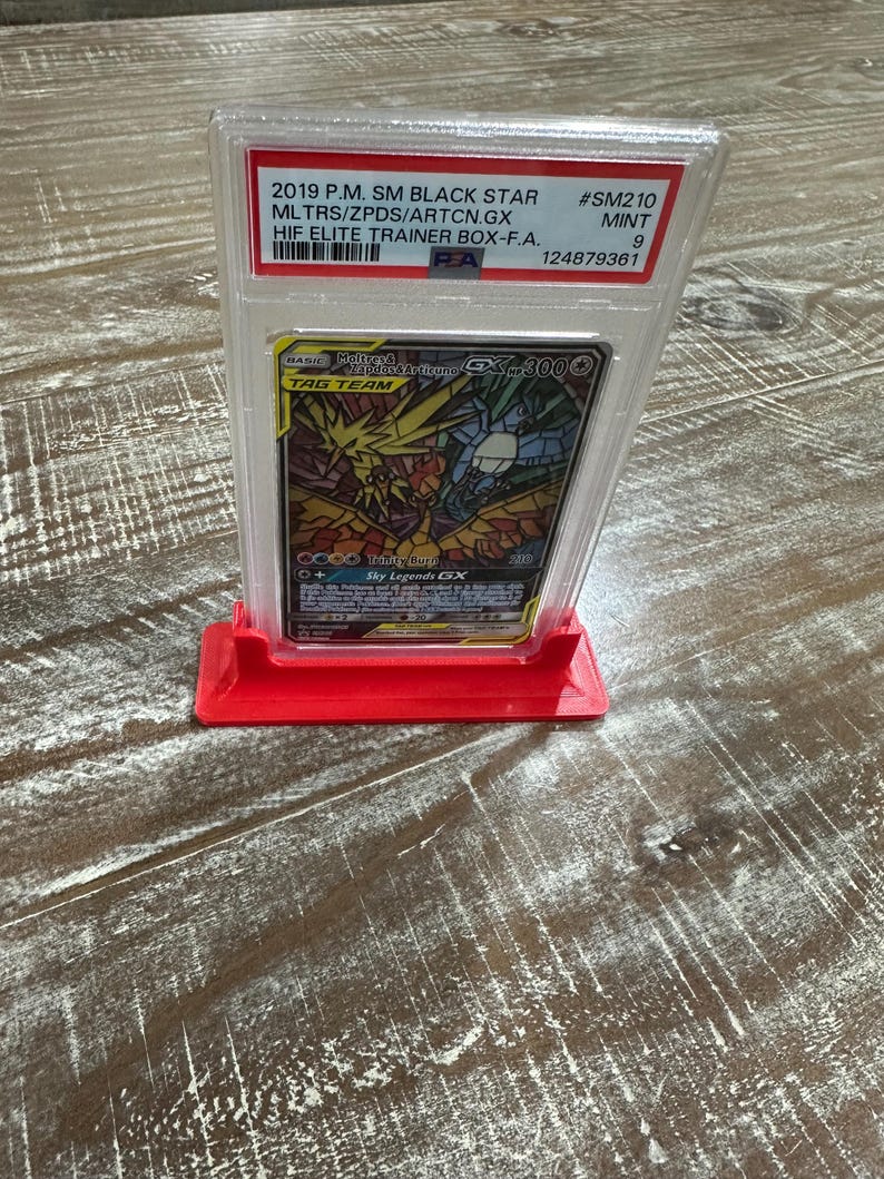 May include: A graded Pok&eacute;mon card in a clear plastic case, displayed on a red stand. The card features a colorful illustration of Moltres & Zapdos & Articuno GX. Text on the case indicates it's a 2019 P.M. SM Black Star card.