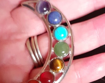 May include: A silver crescent moon pendant necklace with seven colorful gemstones. The stones are arranged in a rainbow pattern, starting with amethyst, then lapis lazuli, turquoise, green aventurine, tiger's eye, carnelian, and red jasper.