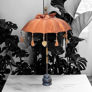 Pode incluir: Um pequeno guarda-chuva de seda laranja com detalhes dourados e borlas. O guarda-chuva está em um suporte preto com uma base redonda.