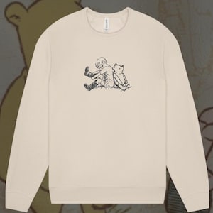 Könnte beinhalten: Cremefarbenes Sweatshirt mit einem schwarzen, gestickten Design eines Jungen und eines Bären. Das Sweatshirt hat lange Ärmel und einen Rundhalsausschnitt. Der Text auf dem Bild lautet "Embroidered Vintage Design Unisex Sizing".