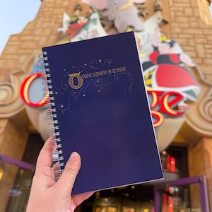 May include: A dark blue spiral-bound notebook with the words "Once Upon a Time" in gold script. The cover features a decorative "O" and star accents. The background includes a building facade with cartoon characters.