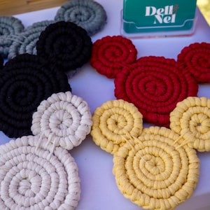 May include: A collection of handmade macrame coasters in the shape of a famous mouse. The coasters are in various colors, including red, yellow, black, white, and gray. A teal card with the text "Dell Nel" is visible in the background.