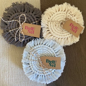 May include: Three handmade macrame coasters in gray, cream, and light blue, each with a fringed edge. Each coaster is tied with twine and has a small tag that reads "Dell & Nel". The coasters are arranged on a textured, two-tone background.