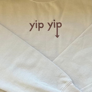 Könnte beinhalten: Ein hellbeiger Pullover mit einem braunen gestickten Design, das "yip yip" mit einem nach unten zeigenden Pfeil sagt.