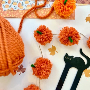 Könnte beinhalten: Nahaufnahme eines herbstlichen Bastelprojekts. Es zeigt eine Girlande aus orangefarbenen Garn-Pompons mit grünen Stielen, einen Knäuel orangefarbenen Garns und einen schwarzen katzenförmigen Lesezeichen. Dekorpapier mit Kürbissen und Blättern.