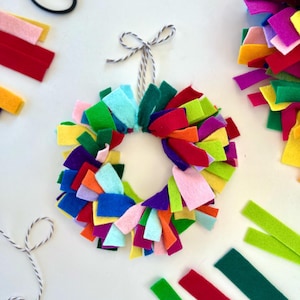 Peut inclure: Une couronne en feutre colorée avec un nœud en ficelle blanche. La couronne est faite de bandes de feutre de différentes couleurs, notamment rouge, orange, jaune, vert, bleu, violet et rose. La couronne est suspendue à une ficelle blanche.