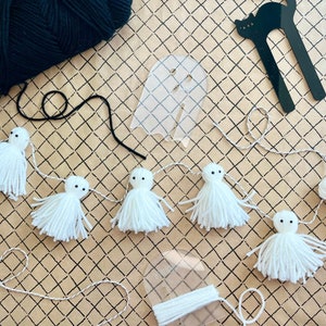 Pode incluir: Uma guirlanda de Halloween feita à mão com fantasmas de fio branco enfiados em um cordão branco. Os fantasmas têm olhos e braços pretos. Uma bola de fio preto, um modelo em forma de fantasma e uma ferramenta de artesanato em forma de gato preto também são visíveis.
