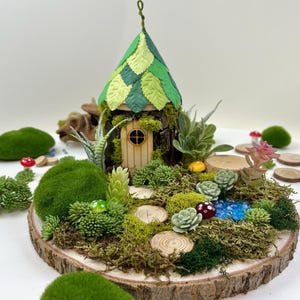 Puede incluir: Una casa de hadas en miniatura con un techo de hojas verdes, rodeada de musgo, suculentas y peque&ntilde;os troncos de madera sobre una rodaja de madera.