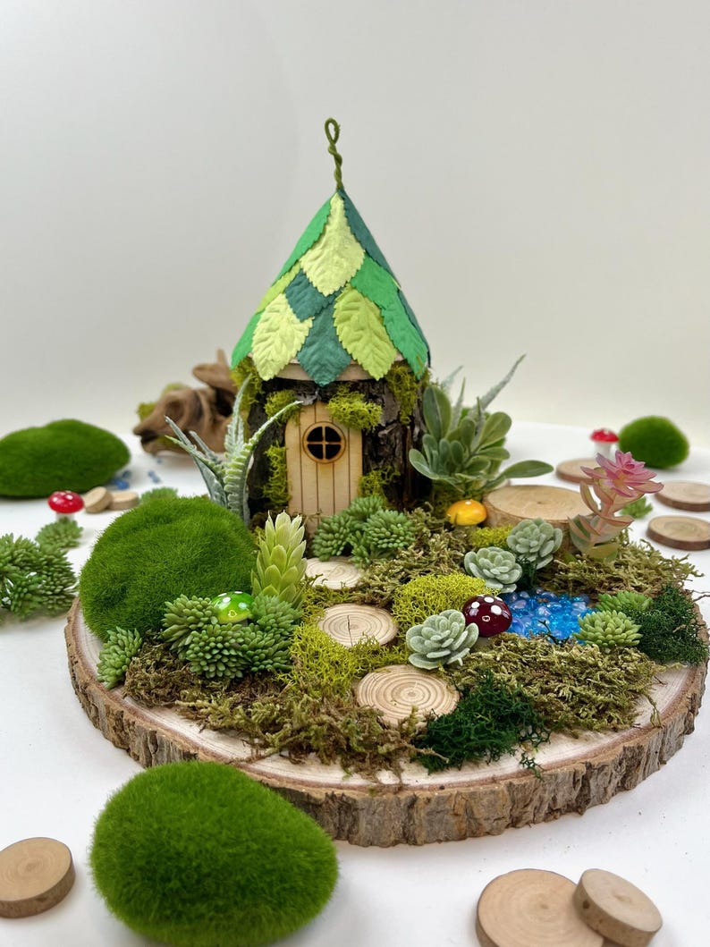 Puede incluir: Una casa de hadas en miniatura con un techo de hojas verdes, rodeada de musgo, suculentas y peque&ntilde;os troncos de madera sobre una rodaja de madera.
