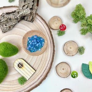 Puede incluir: Una casa de hadas de madera con una peque&ntilde;a puerta y una ventana, rodeada de &aacute;rboles en miniatura, musgo y un cuenco de cuentas de vidrio azul. La escena est&aacute; ambientada en una rodaja de madera.