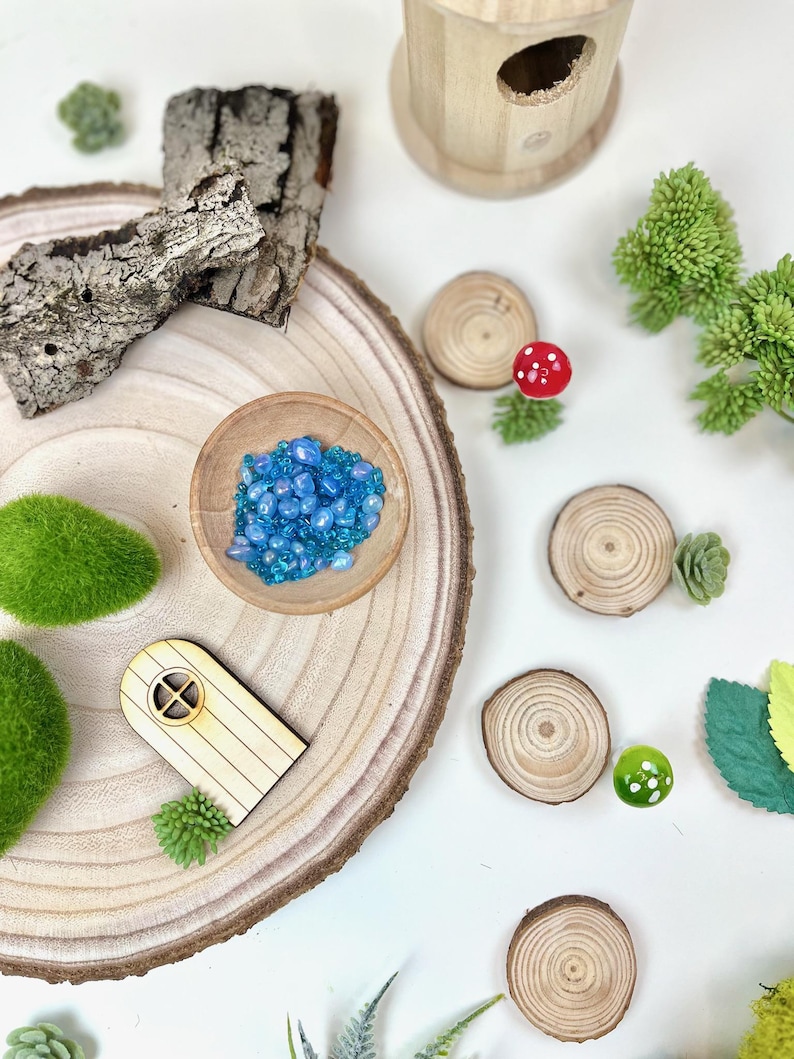 Puede incluir: Una casa de hadas de madera con una peque&ntilde;a puerta y una ventana, rodeada de &aacute;rboles en miniatura, musgo y un cuenco de cuentas de vidrio azul. La escena est&aacute; ambientada en una rodaja de madera.
