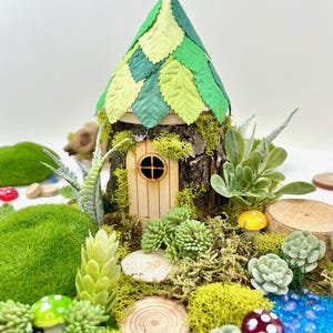 Puede incluir: Una casa de hadas en miniatura hecha de madera y hojas verdes, con una peque&ntilde;a puerta de madera y una ventana. La casa est&aacute; rodeada de musgo, peque&ntilde;as plantas y rodajas de madera.