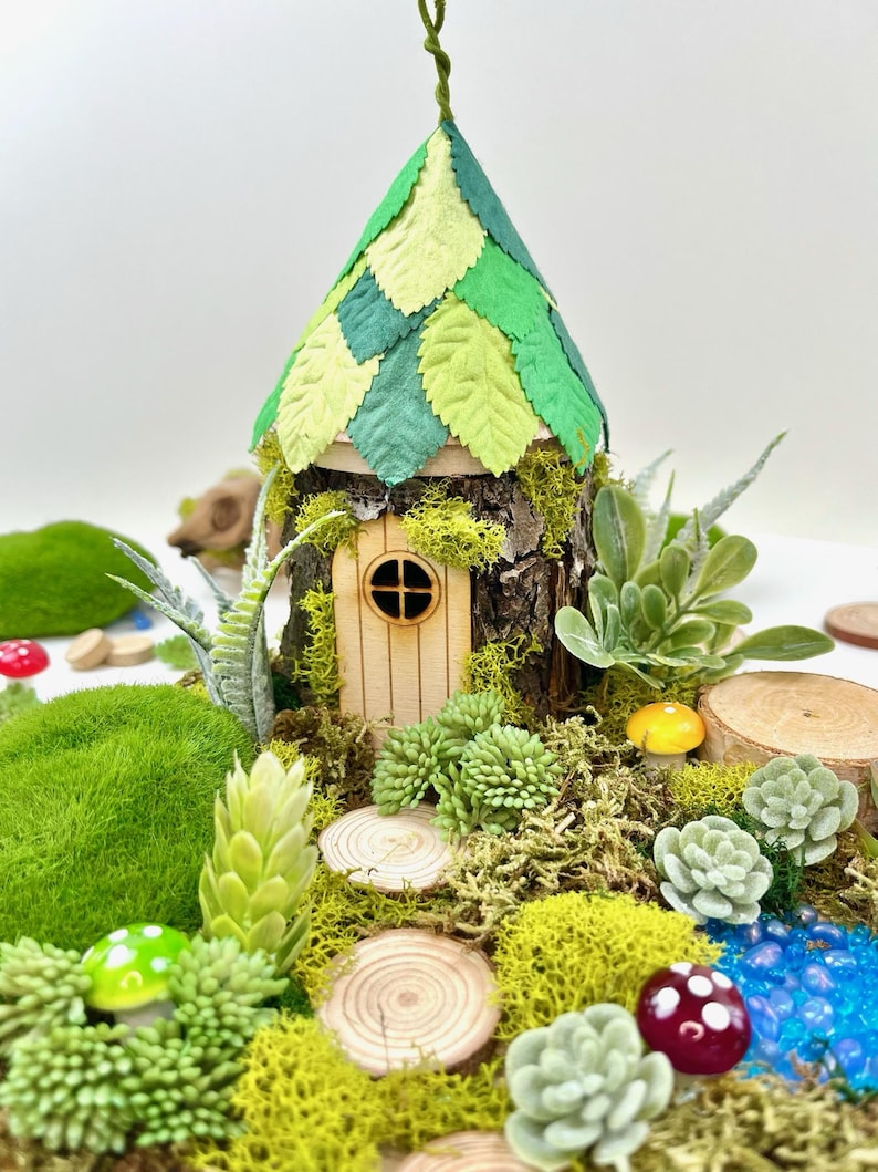 Puede incluir: Una casa de hadas en miniatura hecha de madera y hojas verdes, con una peque&ntilde;a puerta de madera y una ventana. La casa est&aacute; rodeada de musgo, peque&ntilde;as plantas y rodajas de madera.