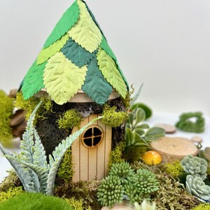 Puede incluir: Una casa de hadas en miniatura hecha de madera y musgo con un techo de fieltro verde en forma de hojas. La casa tiene una peque&ntilde;a puerta y ventana de madera. La casa est&aacute; rodeada de musgo verde y peque&ntilde;as plantas.