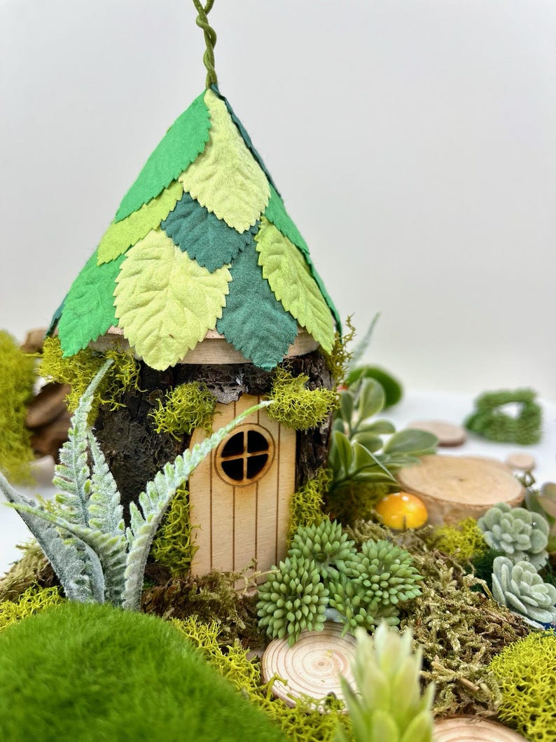 Puede incluir: Una casa de hadas en miniatura hecha de madera y musgo con un techo de fieltro verde en forma de hojas. La casa tiene una peque&ntilde;a puerta y ventana de madera. La casa est&aacute; rodeada de musgo verde y peque&ntilde;as plantas.
