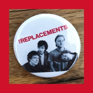 以下が含まれることがあります： 白地に黒と白の写真が印刷されたボタン。バンド名は「The Replacements」で、文字は赤色です。