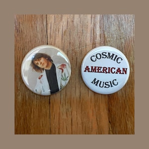 May include: Two round button pins. One pin features a black and white photo of a person with long hair. The other pin is white with black text that reads "COSMIC AMERICAN MUSIC".