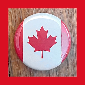 May include: A round button with a white background and a red maple leaf in the centre. The button has a red, white, and red border.