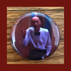 Puede incluir: Un botón con la imagen de un hombre que lleva una camisa blanca con una pajarita negra, haciendo un gesto de pulgar hacia arriba. Sonríe y tiene la cabeza calva.