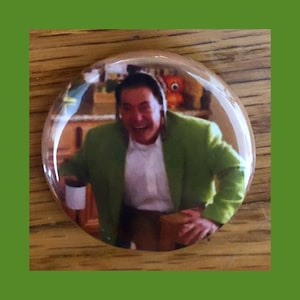 Puede incluir: Un botón con un hombre con una chaqueta verde riendo. Está sosteniendo una taza blanca en su mano izquierda y una caja marrón en su mano derecha. El fondo está borroso y muestra una cocina.