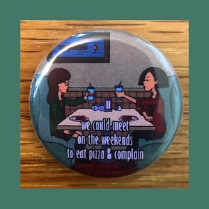 Peut inclure: Un bouton rond avec une image de bande dessinée de deux femmes assises à une table dans un restaurant. Le texte sur le bouton dit "we could meet on the weekends to eat pizza & complain".
