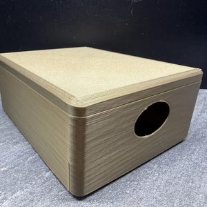 Könnte beinhalten: Eine rechteckige, beige Box mit einer abgerundeten ovalen Öffnung an einer Seite. Die Box hat eine strukturierte Oberfläche und scheint aus einem starren Material zu bestehen. Die Box steht auf einer grauen Oberfläche.