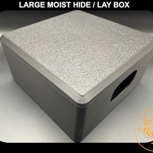 Puede incluir: Una caja rectangular gris para esconderse/poner huevos con una tapa texturizada y un cuerpo gris oscuro. La caja tiene una abertura en forma de asa en un lado. El texto "LARGE MOIST HIDE / LAY BOX" está en la parte superior.