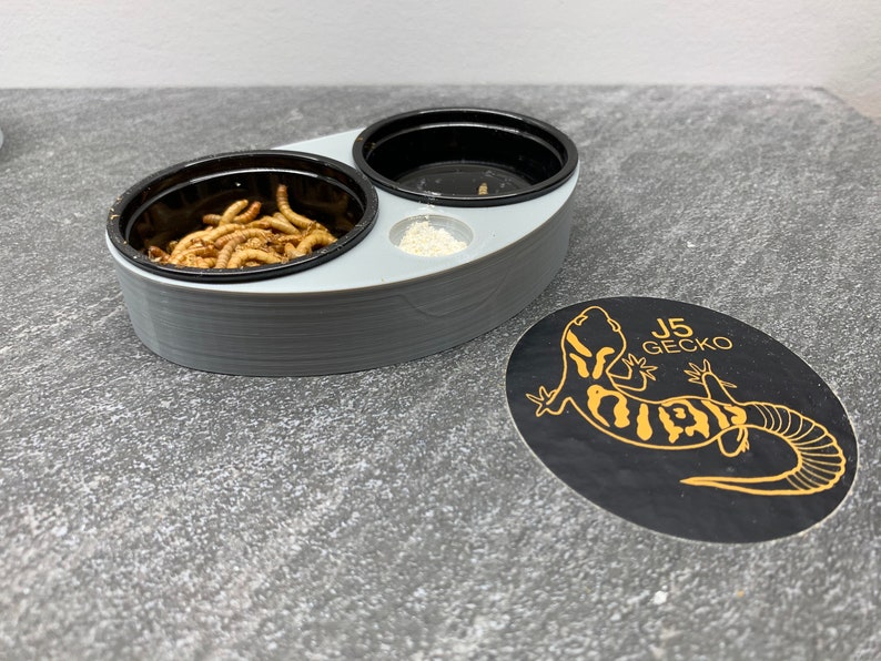 May include: A gray plastic reptile feeding dish with two compartments. One compartment contains dried mealworms, the other is empty. The dish is on a gray textured surface with a black and gold sticker that says "J5 Gecko".