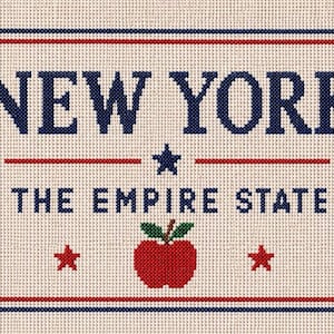May include: A cross-stitch embroidery on beige fabric featuring the text NEW YORK in dark blue, followed by THE EMPIRE STATE. A dark blue star sits between the lines, with a red apple and two small red stars below, all enclosed in a red and blue border.