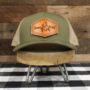 May include: A green and beige trucker hat with a brown leather patch that says "Lone Wolf" and features a wolf howling at the moon.