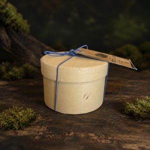 May include: A round, light brown cardboard gift box tied with thin blue twine. A rectangular paper tag with printed text is attached to the top of the box by the blue string. The box has a smooth, matte finish and a simple, cylindrical shape.