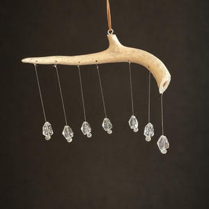 May include: A hanging mobile featuring a single curved, light-colored antler branch suspended by a thin brown cord. Seven clear, faceted crystal-like pendants hang from the antler at varying intervals, each attached by a thin, silver-toned wire.