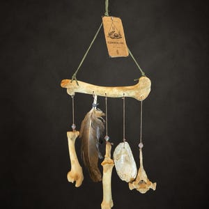 May include: A hanging mobile featuring a large horizontal bone as the main support, suspended by a green cord with a tan tag labeled ELEMENTAL LINE. Five items hang from the bone: two smaller bones, a brown feather, a shell, and a vertebrae.