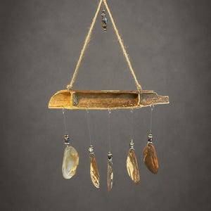 May include: A hanging mobile featuring a horizontal piece of weathered driftwood with a hollow center. Five thin strings hang from the wood, each adorned with a small metallic bead and a translucent, irregularly shaped shell in shades of brown.