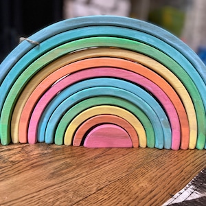 May include: A rainbow stacking toy made of wood with pastel colors. The toy is made of 10 pieces, each a different size and color. The pieces are arranged in a rainbow pattern, with the largest piece at the bottom and the smallest piece at the top.