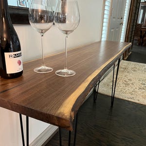 May include: A live edge console table with a dark wood finish and black hairpin legs. A bottle of wine and two wine glasses sit on the table. The table is approximately 60 inches long.