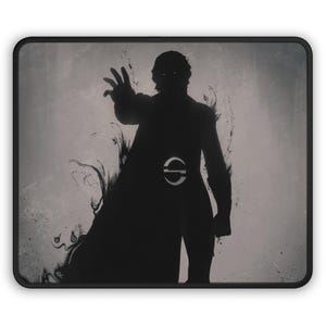 May include: A rectangular mouse pad with a black stitched border featuring a dark silhouette of a figure with glowing eyes and a hand outstretched. The figure has a circular emblem on the chest and is surrounded by swirling smoke-like effects.