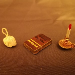 Könnte beinhalten: Ein Miniaturbuch mit dem Titel "Grimm's Fairy Tales" mit braunem Einband und goldenen Akzenten, einer weißen gehäkelten Glocke und einem goldenen Kerzenleuchter mit einer roten Kerze.