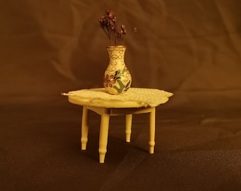 Puede incluir: Un pequeño jarrón blanco con estampado floral con flores secas se encuentra sobre una mesa redonda blanca de cuatro patas. La mesa y el jarrón están sobre una superficie marrón.
