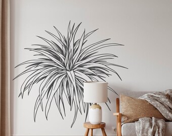 May include: A black and white line drawing of a large plant with long, thin leaves. The drawing is on a white wall in a living room setting. A wooden side table with a lamp and a couch with a throw blanket are visible in the background.