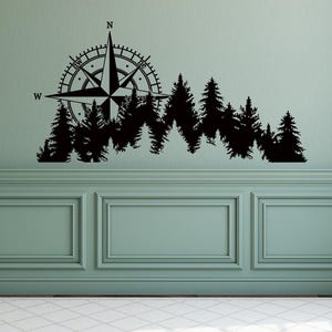 Könnte beinhalten: Schwarzer Wandaufkleber mit einem Kompassrosen-Design über einer Waldsilhouette. Der Kompass zeigt nach Norden, Westen und Nordwesten. Die Waldsilhouette umfasst mehrere Kiefern. Der Aufkleber ist an einer grünen Wand angebracht.