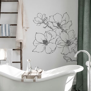 May include: A white bathtub with a wooden tray and silver glasses sits in a bathroom with a white wall featuring a black and white floral decal. A silver freestanding faucet is next to the tub.