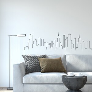 K&ouml;nnte beinhalten: Ein wei&szlig;es Sofa mit zwei Kissen, einem grauen und einem braunen, steht vor einer wei&szlig;en Wand mit einer schwarzen Umrisszeichnung einer Skyline. Eine schwarze Stehlampe steht links vom Sofa. Ein kleiner Holztisch mit zwei wei&szlig;en Tassen steht vor dem Sofa.