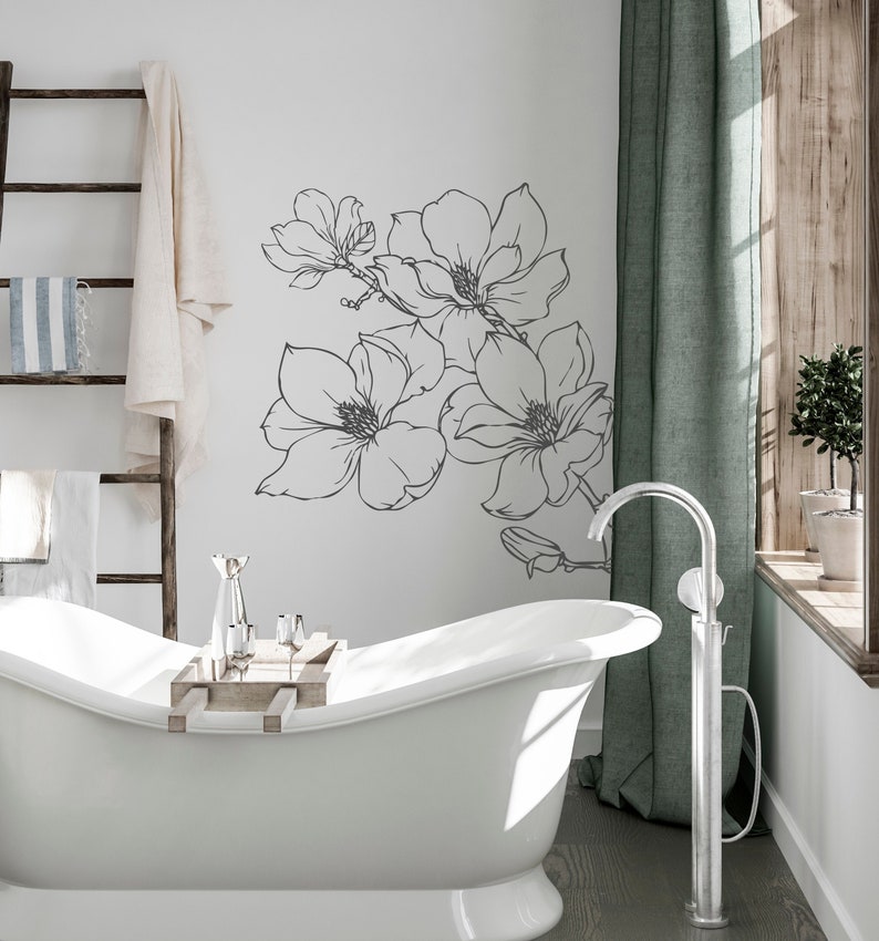 May include: A white bathtub with a wooden tray and silver glasses sits in a bathroom with a white wall featuring a black and white floral decal. A silver freestanding faucet is next to the tub.