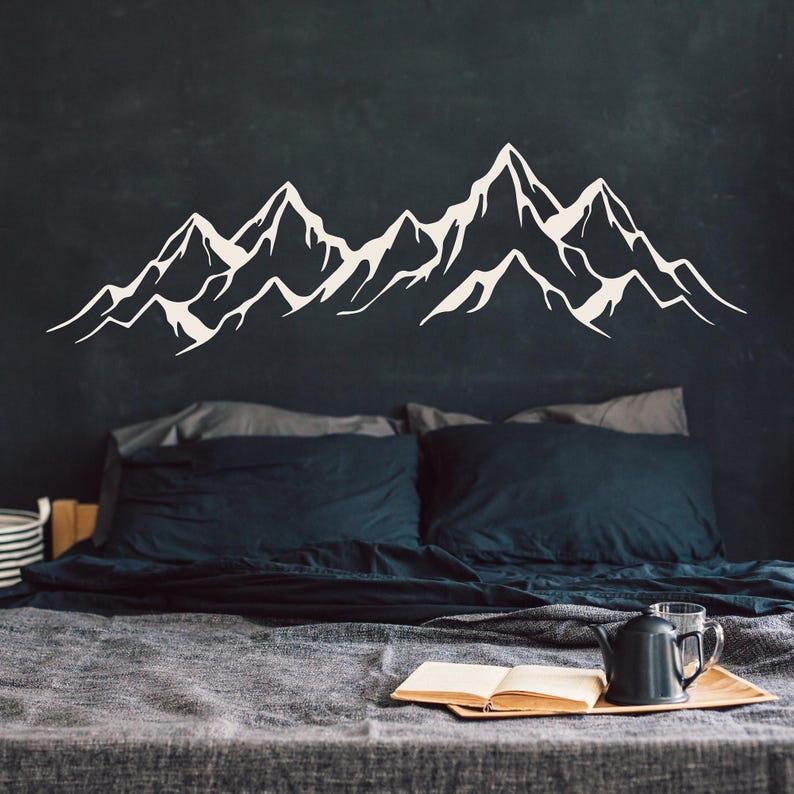 May include: A white mountain range silhouette wall decal on a dark gray wall above a bed with dark gray bedding and a book on a wooden tray.