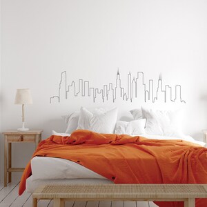 K&ouml;nnte beinhalten: Ein wei&szlig;es Bett mit einem wei&szlig;en Kopfteil und einer orangefarbenen Decke. Das Kopfteil zeigt eine schwarze Umrandung einer Stadtlandschaft.