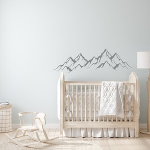May include: A white crib with a white quilt and a gray mountain range wall decal in a nursery setting.